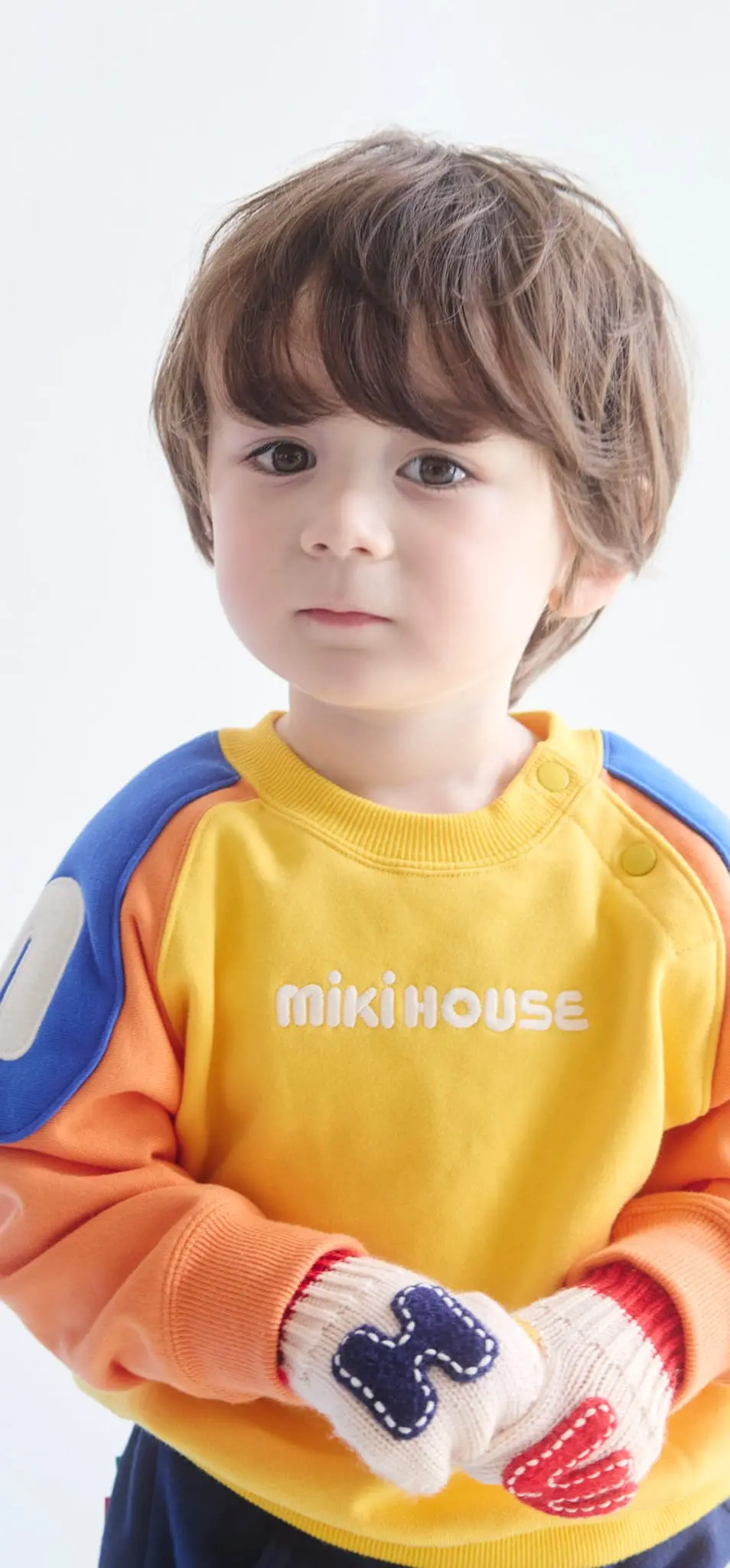 青いトレーナーを着て元気にポーズをとる、笑顔の子どもが白い壁の前に立っている